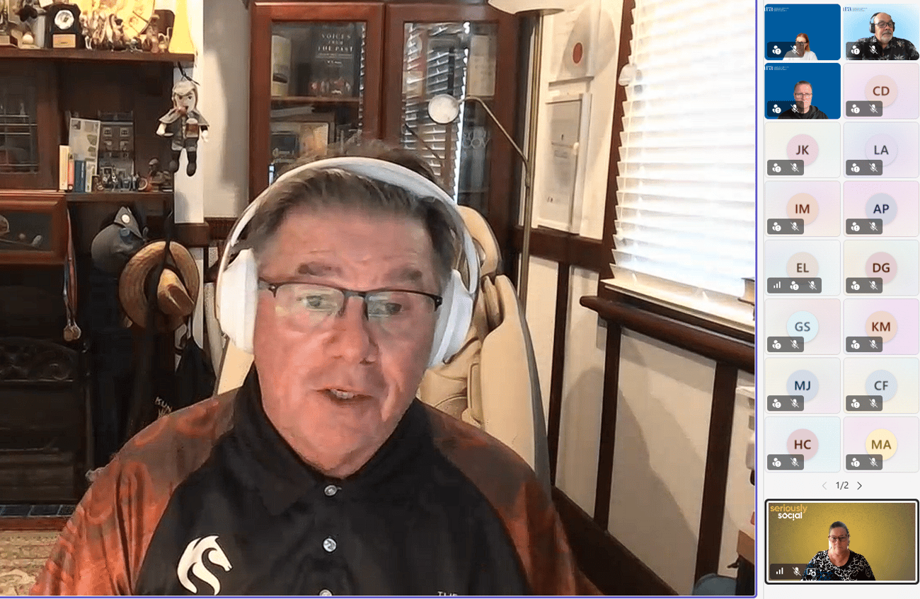 Professor John Maynard wears a dark polo shirt with Indigenous patterns on the sleeves, and sits in front of a bookshelf and window as he presents to an audience via webinar. He is wearing white headphones.