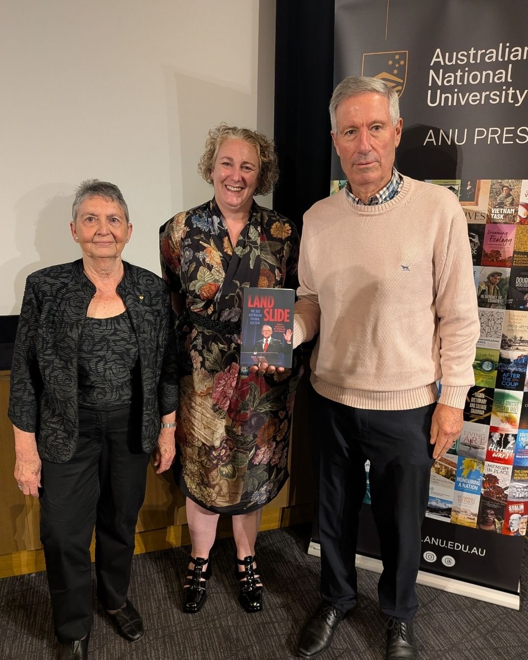 Two women and a man stand in front of an ANU banner. The man holds a copy of the book "Landslide: the 2025 Australian Federal Election"