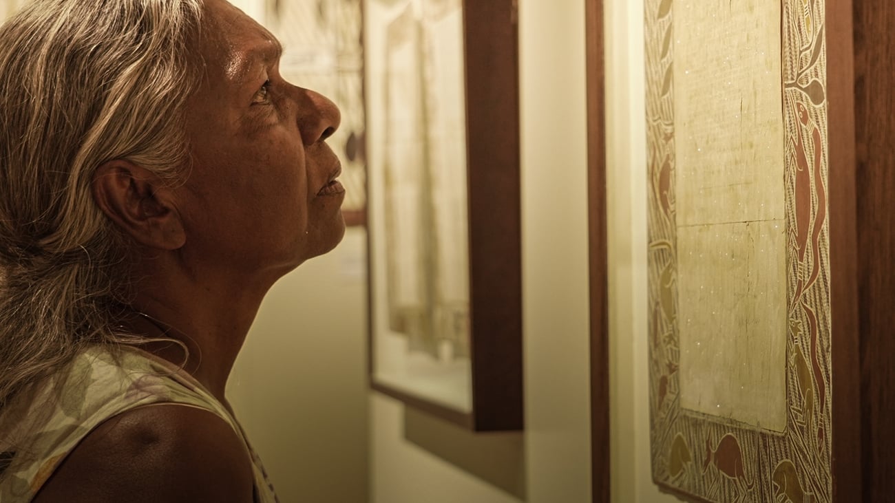 A promotional image for the One Mind, One Heart film screening and Q&A session. The image features a woman with brown skin and long grey hair looking at a framed piece of writing. There are Aboriginal art designs around the frame. 
